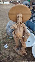 Full view of hand carved wooden cowboy statue with large sombrero, bottle in left hand, and three-finger shooter gesture with right hand, standing outdoors with background of miscellaneous items.