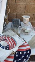 Photo showing various home items on a white surface, including American flag chip and dip tray, crystal-like swirl bowl containing some clips, floral ceramic vase, small metal frame, clear glass dish, and round textured glass plate.