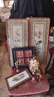 Two framed silk embroidered wall hangings with floral and bird designs, painted picture in frame, geisha doll, miniature vase with hat pins, and two wooden carved figurines arranged on a chair.