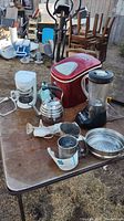 Full view of kitchen appliances and accessories on table outdoors showing red Igloo ice maker, white Mr. Coffee drip coffee maker, stainless steel teapot, black Hamilton Beach blender, white handheld mixer, metal sifter, steamer insert, and strainer.
