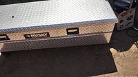 Photo of aluminum Husky toolbox with diamond plate pattern, key lock on lid, shown closed and placed on ground.