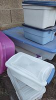 Set of seven small plastic totes stacked outside against a brick wall, various colors including white, blue, purple, and clear, some with lids.