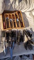 Photo showing stainless steel flatware pieces including forks, knives, and spoons arranged in a wooden tray, with additional kitchen knives and utensils to the side on fabric surface.