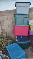 Six stacked Rubbermaid storage totes in various colors against a brick wall, one blue tote is on the ground in front.