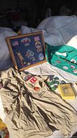 Photo showing vintage Boy Scout tan shirt with patches, green vest with patches, patch display case, scout pins, and handbook on white surface.