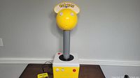 Front view of the yellow spherical joystick head with Pac-Man logo on semicircular sign above, mounted on tall gray shaft and yellow base with buttons and foot pedal.