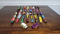 Overview showing 50 assorted toy cars arranged in rows on a wooden table, various types and colors.