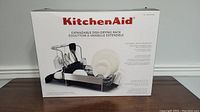 Box showing KitchenAid brand and product image of the expandable dish drying rack with utensils and dishes.