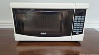 White RCA microwave with front door and visible RCA logo, placed on a brown wooden surface against a light wall.