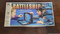 Front of the vintage Battleship game box showing children playing the game with plastic vertical grids and ships, logo and brand MB Milton Bradley, and game description.