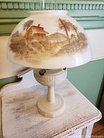 Front view of entire lamp showing painted rural landscape shade and milk glass base on distressed white table