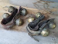 Two sets of antique brass horse bells on leather straps placed on a rough wood surface, showing overall condition and metal patina.