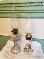 Pair of antique oil lamps shown side by side on a small table against a pale green and white wall; one lamp is clear glass, the other decorated with painted flowers, both with chimneys and metal burners.