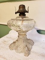 Full view of the antique oil lamp with clear pressed glass and detailed feather and scroll pattern, metal burner on top without chimney.