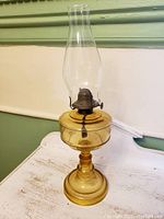 Full view of amber glass oil lamp on white wooden surface with green wall background showing lamp's overall condition and structure.