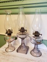 Three early 1900s coal oil lamps displayed side by side on a white surface against a green wall, showing overall condition and proportions.