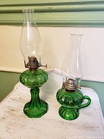 Pair of green glass oil lamps with leaf decorative patterns on a white surface against a cream wall with green trim. Taller lamp on left, smaller handled lamp on right.