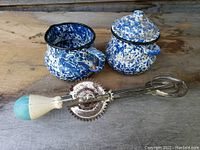 Blue and white spatterware creamer and sugar container with lid and a vintage hand beater with wooden painted handle placed on a wooden surface.
