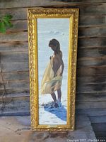 Full view of the vertical elongated oil on canvas painting framed in ornate golden floral carved frame showing a young girl holding cloth standing in shallow water at the beach.