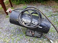 View showing heater body with hose and propane regulator coiled on top on gravel surface outdoors.