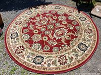 Full view of the round rug laid flat on gravel showing red field and ivory floral border under natural light