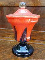 Full front view of a tall orange and black decorated glass covered dish with a pedestal base and clear glass knob handle on top.