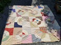 Full view of a vintage handmade quilt laid out on gravel showing multicolor circular block patchwork pattern and visible wear/fading.
