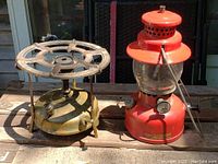 Photo showing vintage brass stove and red lantern side by side on wooden surface under natural light.