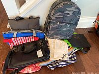 Photo showing multiple bags including a camouflage backpack, various fabric totes and a black satchel handbag.