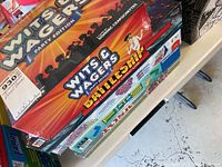 Stack of board games showing Wits & Wagers Party Edition on top, Battleship below and others underneath on shelf.