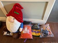 Photo showing large plush gnome toy with red pointed hat and several Halloween costumes in plastic bags on floor near baseboard.
