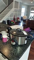 Large stainless steel crockpot with glass lid sitting on kitchen counter with coffee cups around it.
