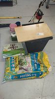 First photo showing large pot, bags of potting mix, wooden board, green plastic container, and several small plastic pots.