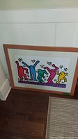 Framed print showing colorful human shapes by Keith Haring leaning against wall on floor.