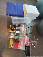 Two stacked plastic storage bins with visible multi-colored folders. On top are a white binder and blue zippered binder. Assorted office supplies and electronics scattered around including cables and digital frame.