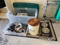 Photo showing a white electric steamer inside a clear plastic bin, a bin of miscellaneous cords, a cream-colored pet food bin with wooden lid, two pet food dishes on a black stand, and an area rug beneath them.