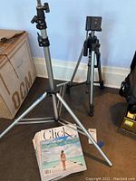 Two silver-tone tripods and a stack of Click photography magazines on floor next to box and wall