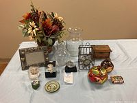 Photo of assorted decorative items on a white cloth including the faux flower arrangement, family picture frame, glass vases, metal fruit basket, trinket boxes, and decorative items.