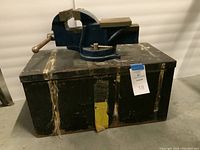 Front and top view of a large heavy-duty blue woodworking vise with wood jaw covers mounted on a worn black toolbox, showing scratches and tape on the toolbox surface.