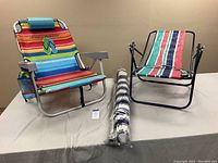 Two beach chairs and a wrapped umbrella on a beige table. Tommy Bahama chair is colorful with carrying straps; second chair is smaller with striped fabric.