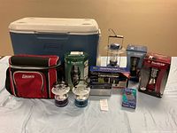 Wide view of lot showing large blue and white Coleman cooler, Coleman insulated cooler bag, two small decorative lanterns, three boxed SteelBEAM lanterns, solar radio and charger, and Radio Shack boxed pocket radio.