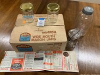 Summary image showing the boxed set of Kerr wide mouth pint mason jars, two jars with gold lids on top of the box, vintage jars and printed canning chart spread out on wooden table.