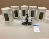 Five white cylindrical canister tins with clear rectangular windows and fitted lids, arranged with a white ceramic salt and pepper shaker set in a dark metal holder with grape leaf decoration.