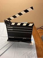 Front view of the black movie film chalk clapboard with sections for Title, Starring, Location, Producer, and Date, sitting on a stand.