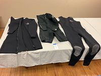 Three wetsuits laid flat on table: ScubaMax 3XL long sleeve short leg, O'Neill XXL short arm and leg wetsuit, ScubaMax 3XL full leg wetsuit with reinforced knees