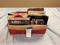 Box containing numerous vintage 45 RPM records in original sleeves from the 60s and 70s, visible labels include Apple Records and A&M Records.