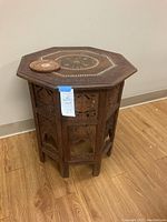 Full view of octagonal vintage collapsible table with detailed carved side panels and decorative inlaid top alongside carved wooden coaster on top.