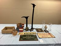 Two cast iron cobbler stands, three Americana flag wooden bins, square woven bread basket, small shaped antique tobacco tin, two glass candleholders, and three vintage metal advertising signs displayed on a table under warm light.
