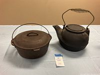 Front view of cast iron kettle with coil handle and Lodge 5 quart Dutch oven with lid side by side on table