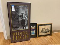Full view of the three vintage wall art pieces on floor against wall: large Riding High photo, Hair The Lovers print and vintage car sketch.